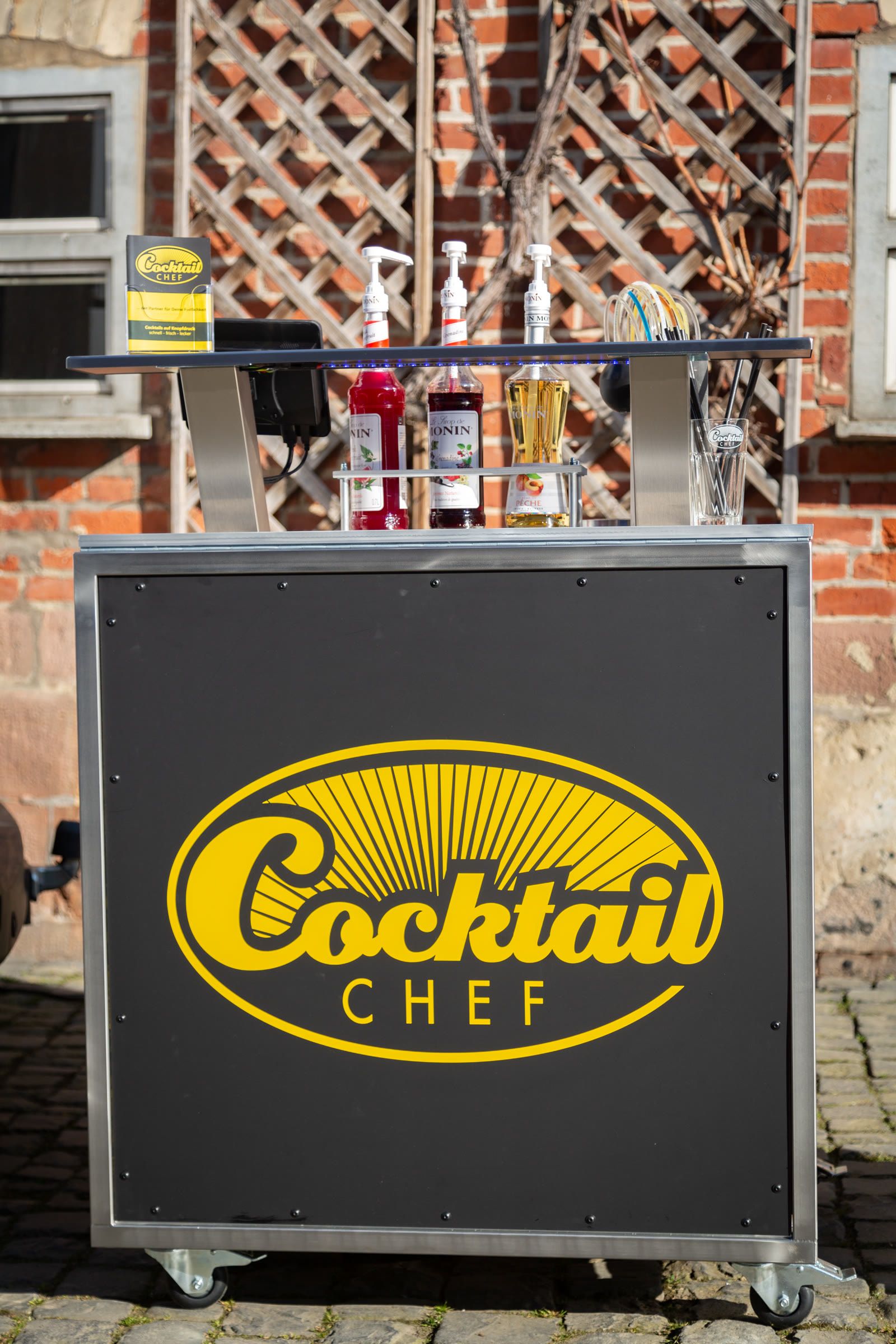 Mobile Cocktailbar mit Cocktailchef-Logo vor einer Backsteinwand